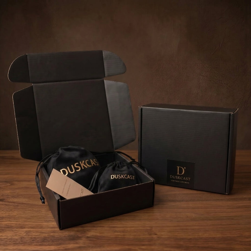 Duskcast vintage-inspired jewelry packaging. Two black velvet pouches inside a matte black mailer box on a dark antique wood table. Old Money aesthetic and dark academia unboxing experience.