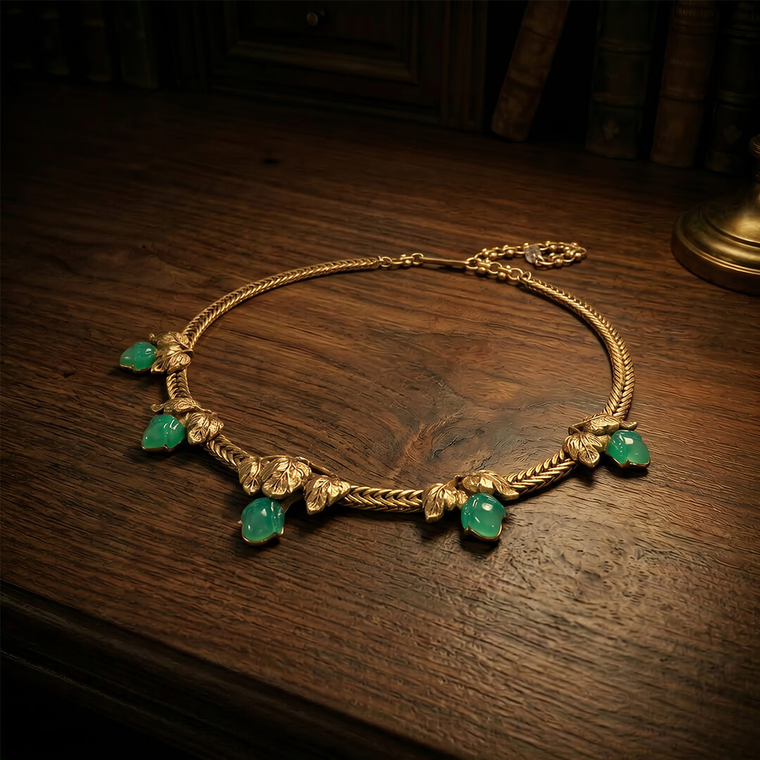 Flat lay view of a heavy gold-tone woven chain necklace featuring botanical leaf motifs and green teardrops.