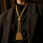 Model wearing a vintage-inspired heavyweight gold braided tassel lariat over a black silk shirt and blazer. 1980s runway revival jewelry and Old Money aesthetic.