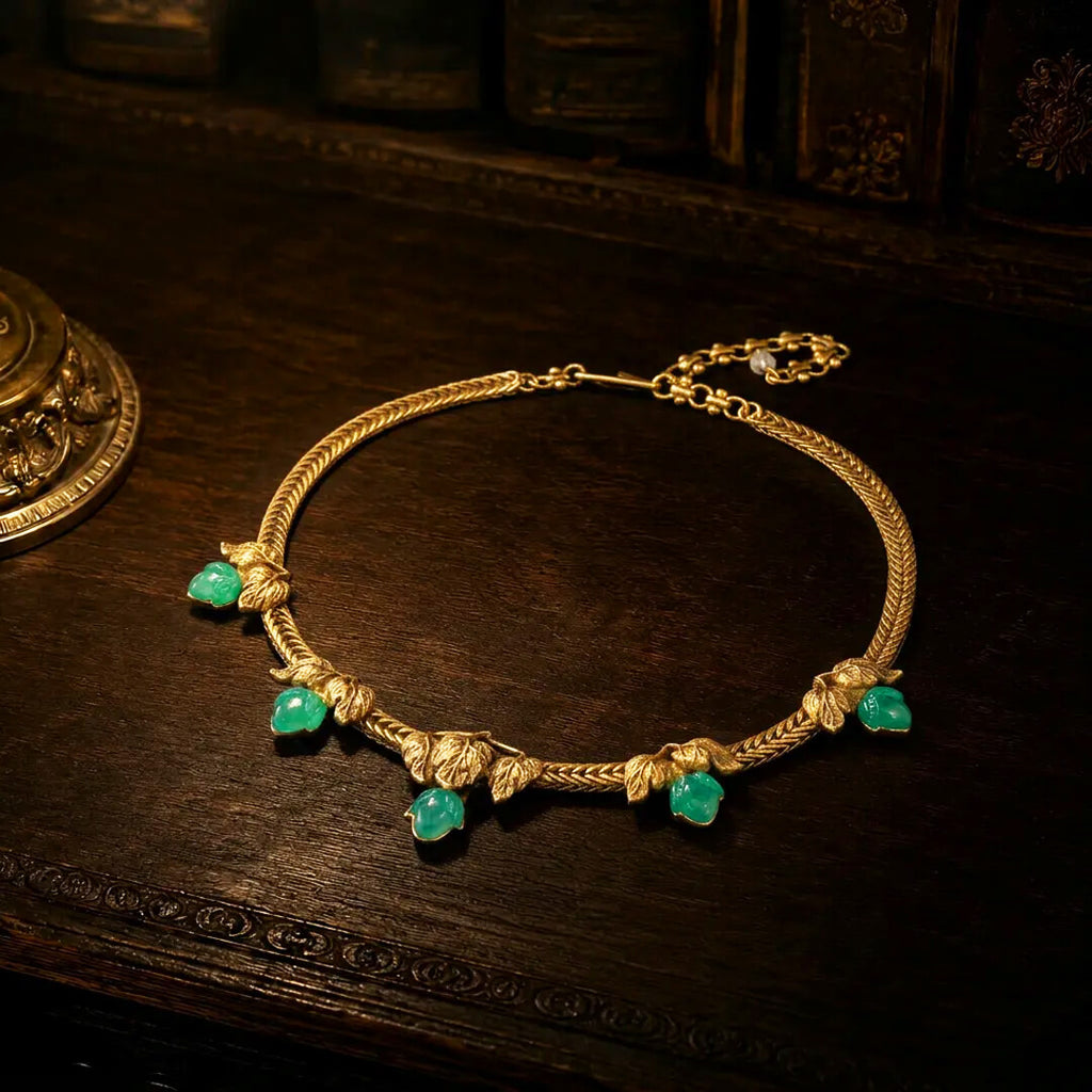 Vintage-inspired gold ivy leaf necklace with green resin stones resting on a dark wood antique desk.