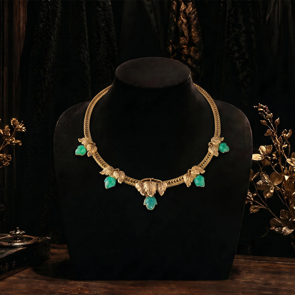 A moody, close-up shot of a vintage-inspired chunky gold collar necklace with five emerald-green cabochon stones resting on a dark, aged, leather-bound antique book.