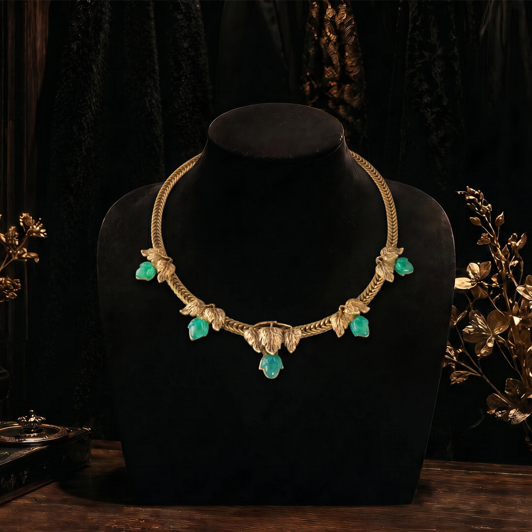 A moody, close-up shot of a vintage-inspired chunky gold collar necklace with five emerald-green cabochon stones resting on a dark, aged, leather-bound antique book.