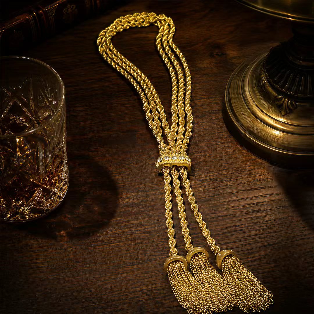 Gilded brass patina tassel necklace laid next to a crystal whiskey glass on a dark wood table. Curated Old Money aesthetic accessories.