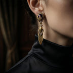 Woman wearing vintage-inspired heavy gold drop earrings with a purple amethyst cabochon, styled with a black silk turtleneck for a dark Old Money aesthetic.