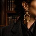 Woman wearing vintage-inspired heavy gold rose chandelier earrings in a dark academia library setting, Old Money aesthetic.