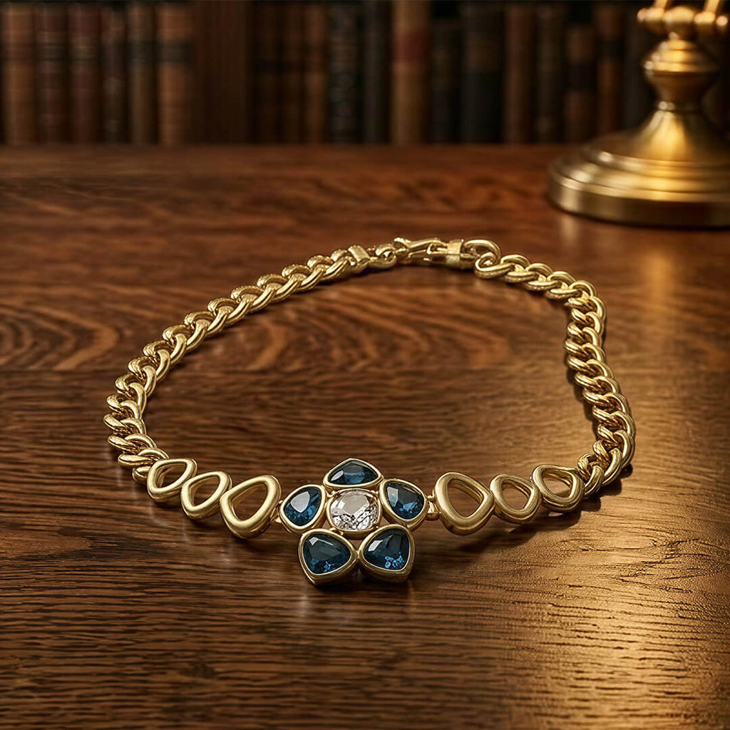 Alt Text: A vintage-inspired 1980s style gold and blue statement necklace laid flat on a rich dark wood antique table next to a brass lamp.
